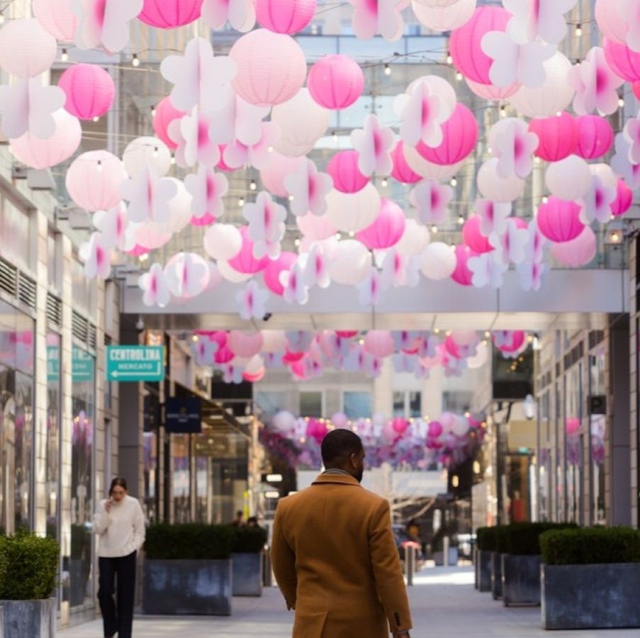 A pedestrian walks through a modern outdoor shopping corridor decorated with hanging pink and white lanterns suspended above the walkway.