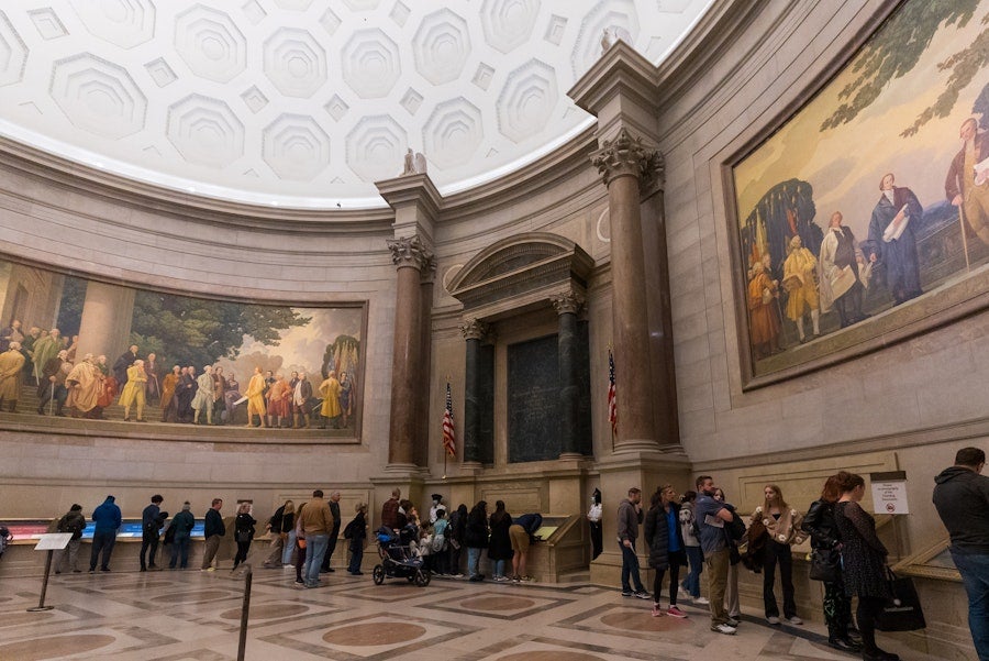 Visitors line up inside the National Archives Rotunda beneath historic murals and a domed ceiling.