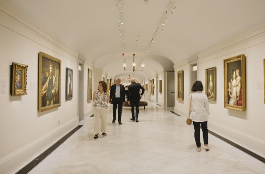 Visitors walk through a long, elegant museum gallery lined with framed paintings on white walls under soft, evenly spaced lighting.
