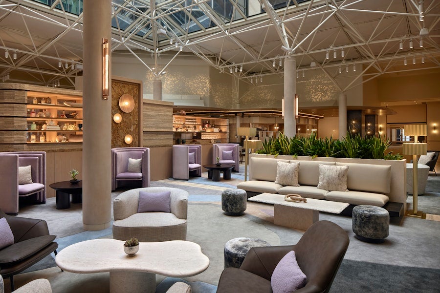A bright, modern hotel lobby with contemporary seating areas, soft neutral tones, indoor greenery, and a high ceiling with geometric skylights.