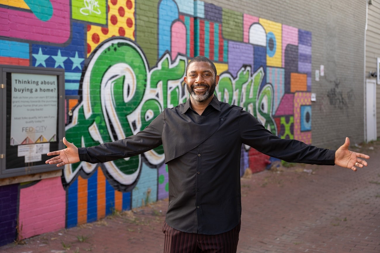 A smiling man stands with his arms open in front of a colorful street mural in Washington, DC.