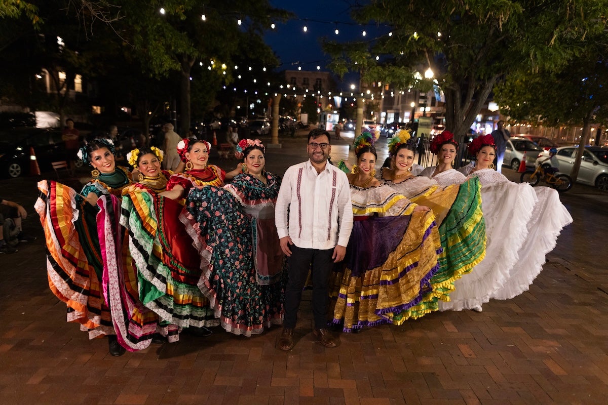 A group of folklórico dancers in colorful traditional dresses pose together outdoors at night during a cultural performance in Washington, DC.