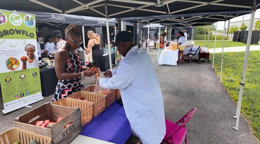 A vendor and customer exchange produce beneath tents at a neighborhood farmers market.