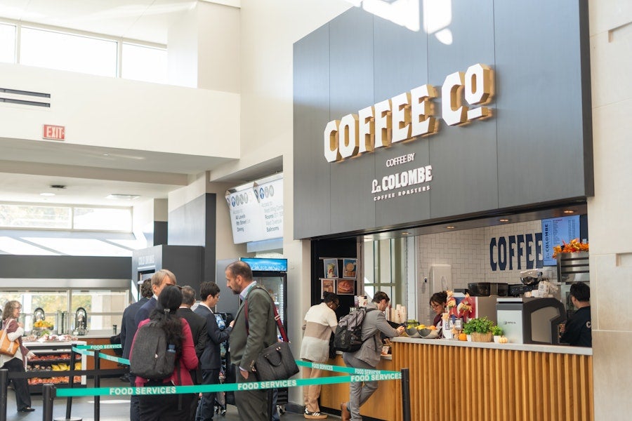People stand in line at a modern indoor coffee kiosk labeled “Coffee Co,” where baristas serve drinks to customers in a bright, open lobby space.