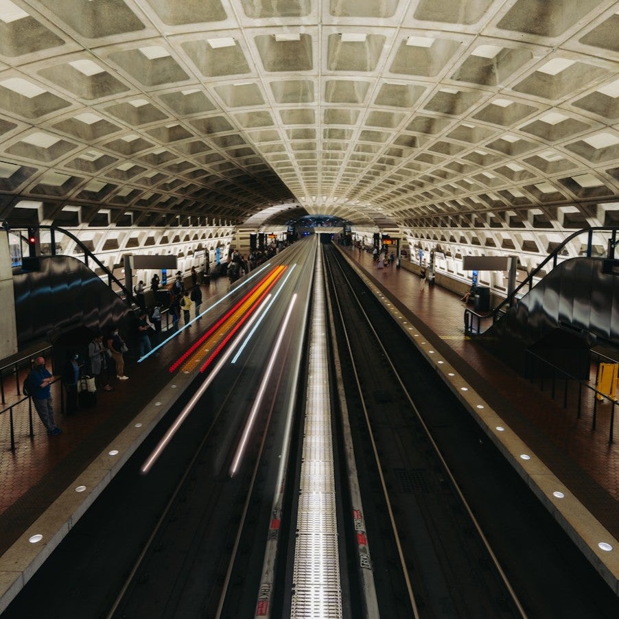 A long-exposure view of a busy underground metro station with a geometric concrete ceiling as a train speeds through the tracks, creating streaks of light while passengers wait on both platforms.