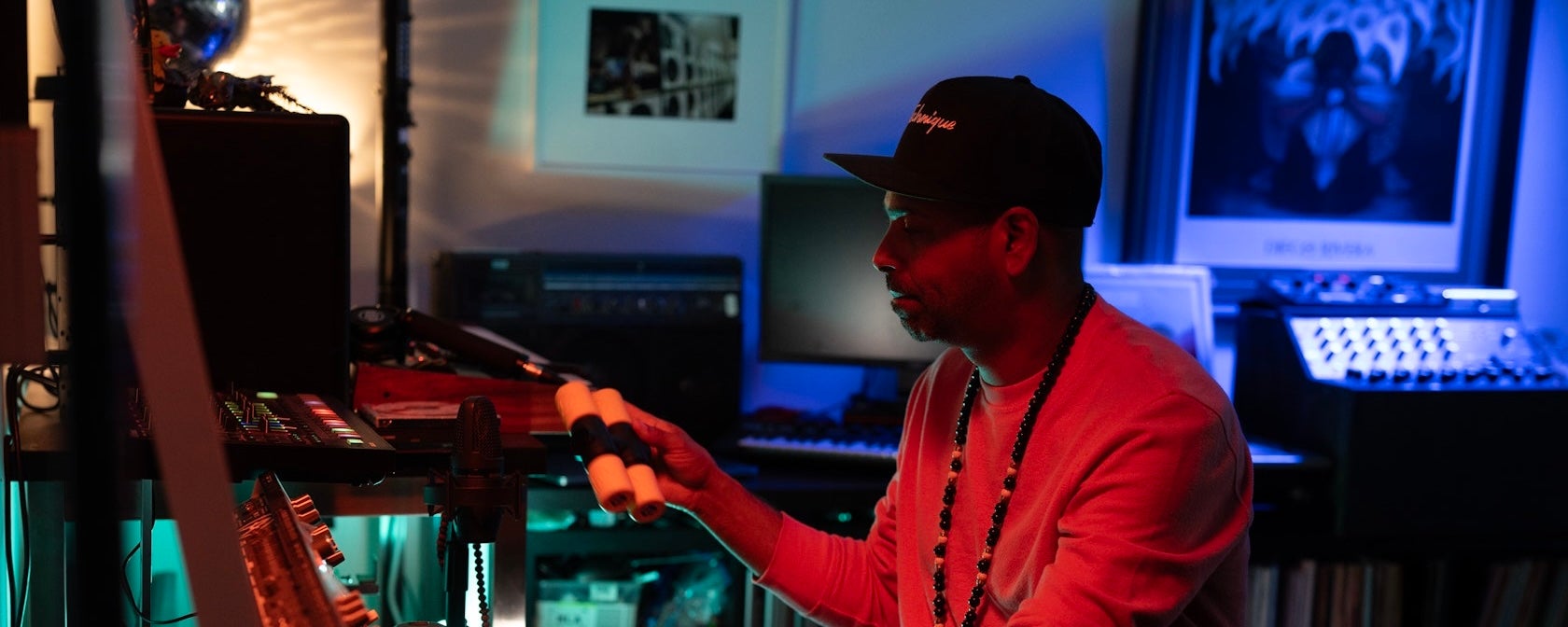 A DJ adjusts equipment in a dimly lit home studio with colorful lighting.