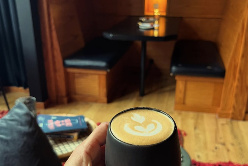 A person holds a cup of latte with leaf-shaped foam art in a cozy café seating area with wooden benches and a small table.