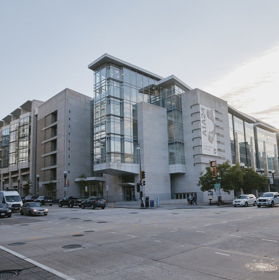 A modern glass-and-concrete museum building on a city corner with cars and pedestrians under a partly cloudy sky.