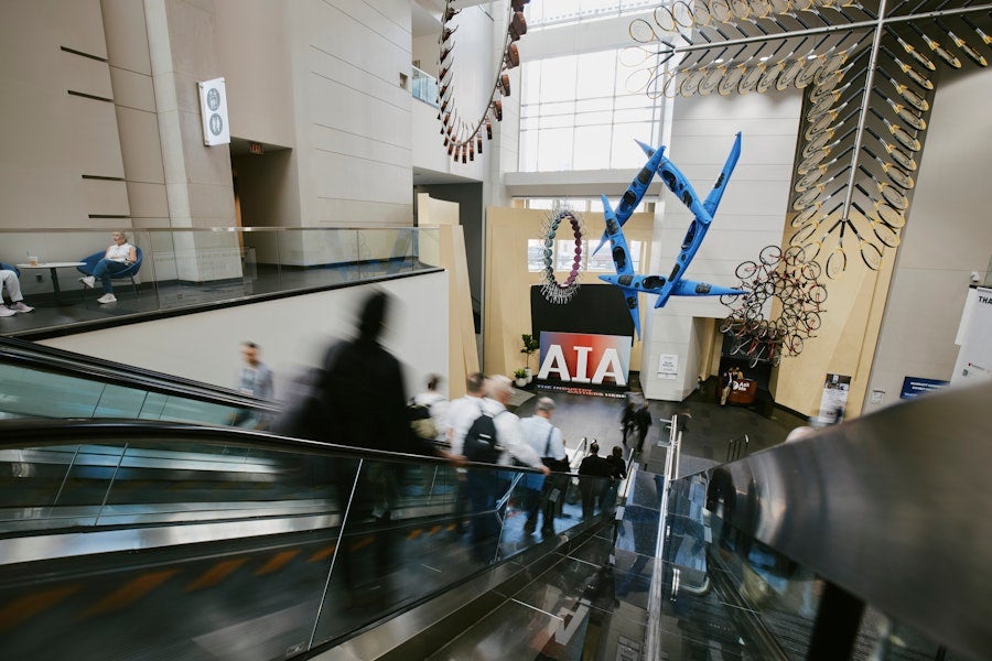 Visitors descend an escalator into a bright museum atrium featuring suspended sculptural installations and a prominent AIA display.