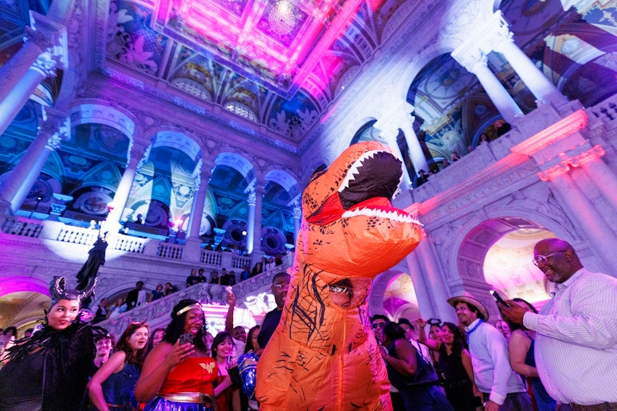 A person in an inflatable T-rex costume dances among a crowd at a colorful, neon-lit event inside an ornate historic hall.
