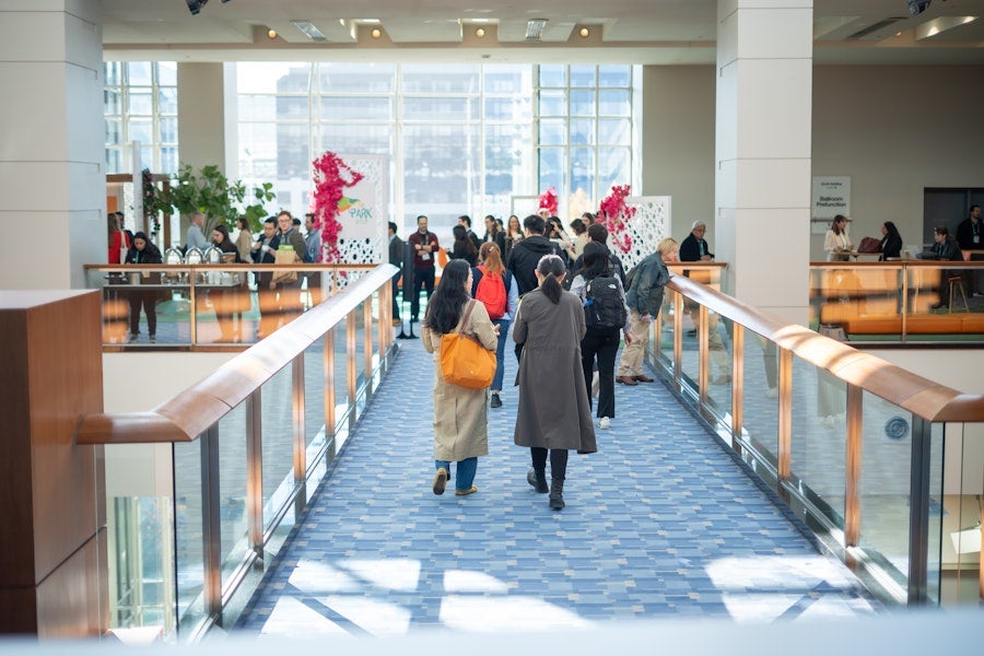 People walk across an indoor skybridge toward a bright gathering area with large windows and event displays.