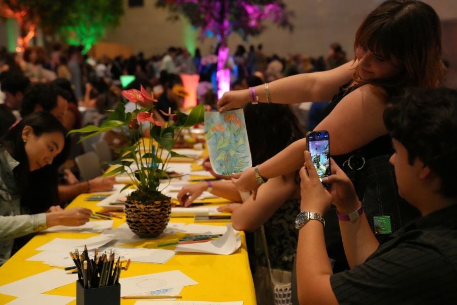 People participating in an art activity at a lively indoor event, with one person holding up a drawing while another takes a photo, surrounded by tables with art supplies and colorful lighting.