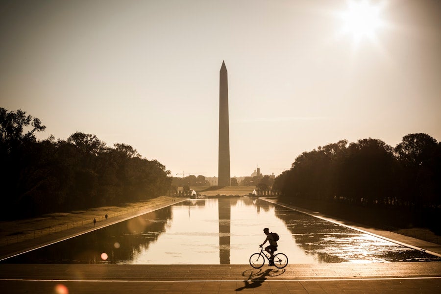 A cyclist rides past the Lincoln Memorial Reflecting Pool at sunrise, with the Washington Monument reflected in the water.