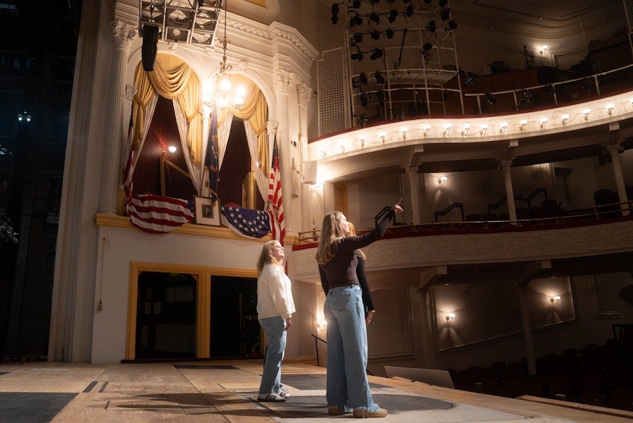 Two people stand on a historic theater stage beneath ornate balconies and American flags, looking up at the lighting above.