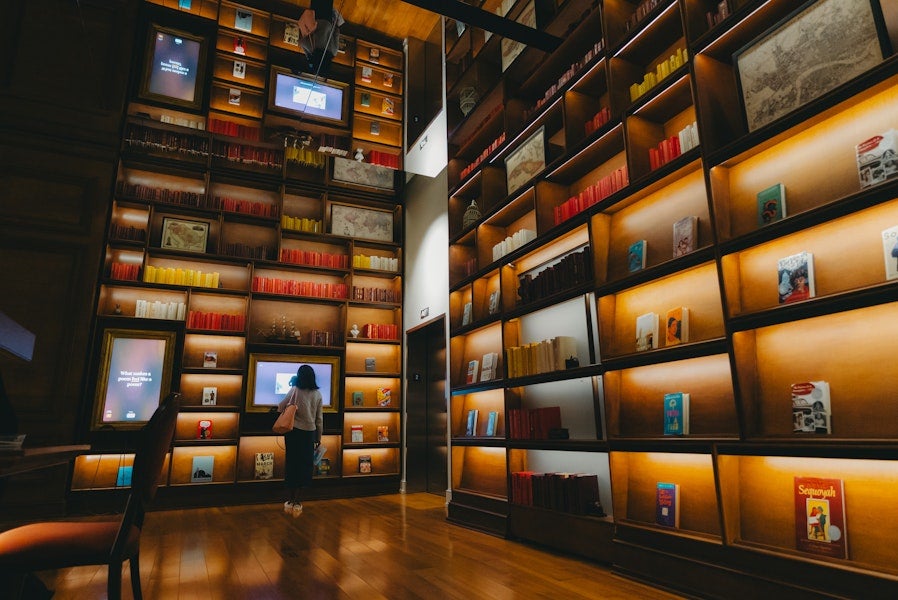 A person explores a warmly lit, floor-to-ceiling bookshelf display with interactive screens in a modern library-like space.