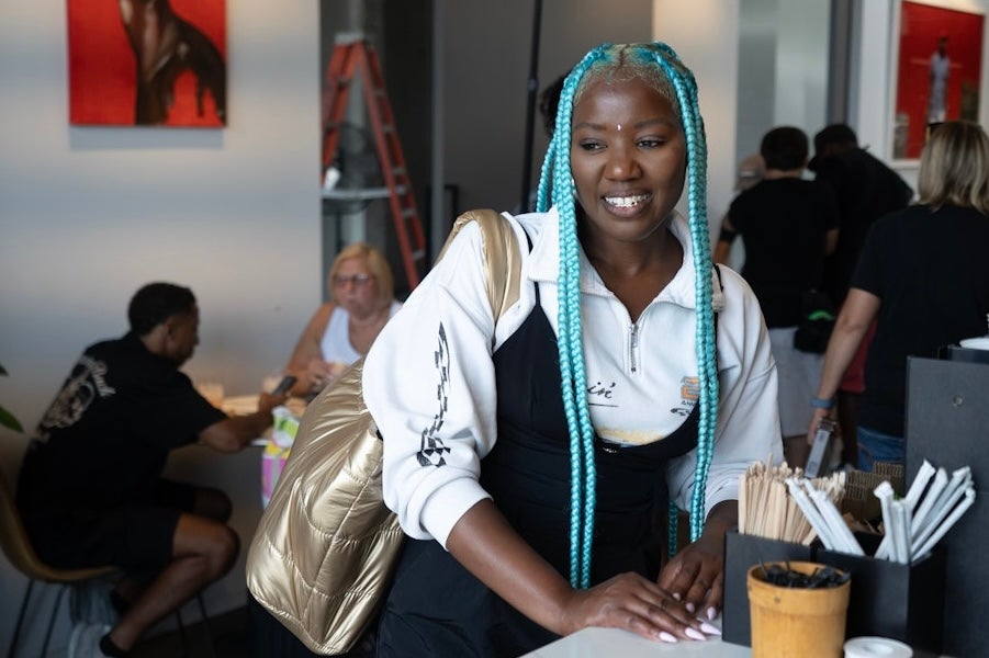 A person with long blue braids smiles while leaning on a café counter with people seated in the background.
