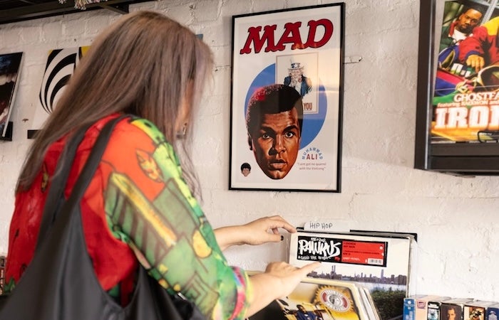 A woman flips through vinyl records in a shop beneath framed posters on the wall.