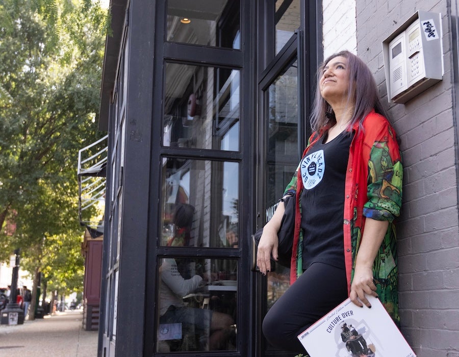 A woman leans against a storefront holding a magazine and looking upward along a tree-lined street.