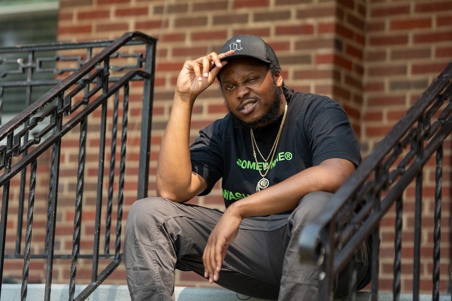 A man in a black T-shirt and cap sits on the steps of a brick building, looking toward the camera.