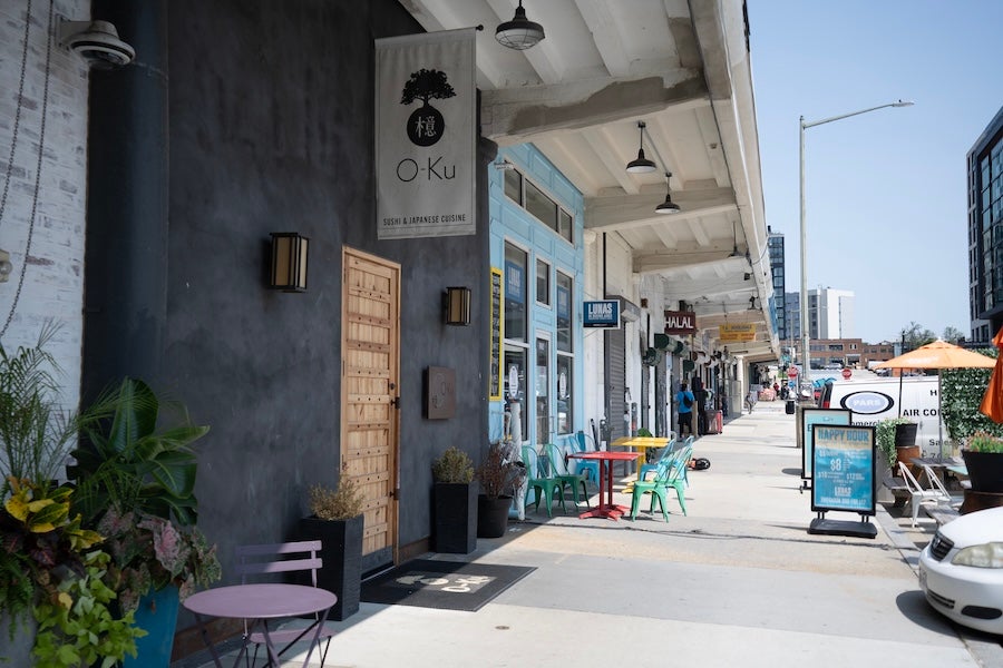 A row of storefronts features outdoor seating and signage outside a restaurant along a sunny sidewalk.
