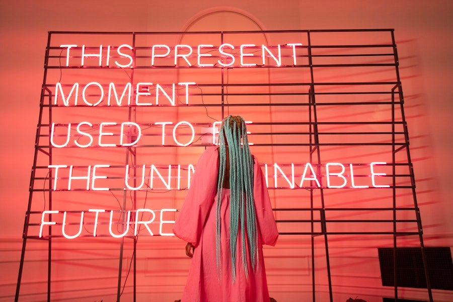 A person with long braids stands facing a glowing neon text installation in a gallery space.
