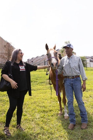 A woman and a man stand beside a horse in a grassy urban setting, smiling and interacting with the animal.