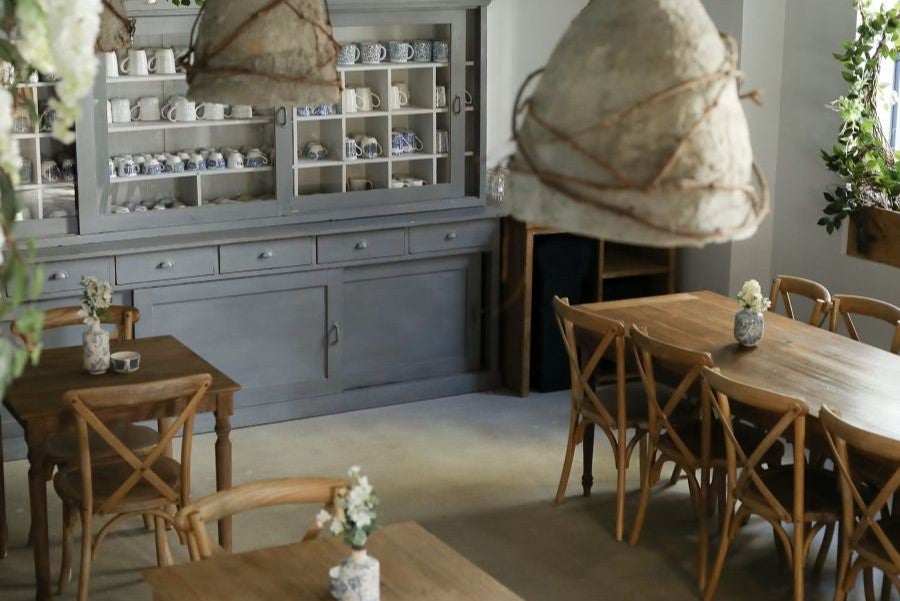 A warm, rustic café interior with wooden tables, cross-back chairs, soft lighting, and a gray hutch filled with neatly arranged mugs.