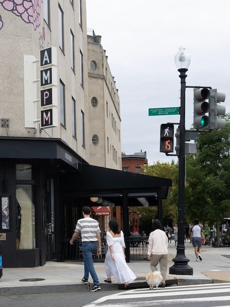 A person in a white dress crosses city street with companions near an Art Deco hotel entrance