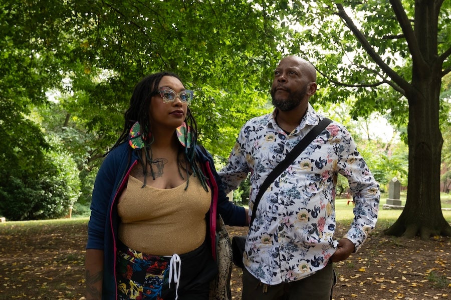 Two people in a leafy park, one holding a phone, both wearing patterned outfits.