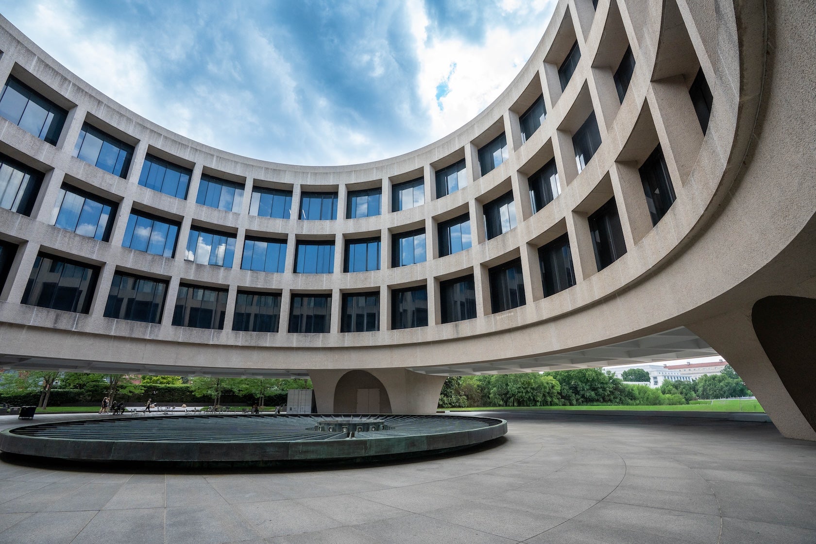Curved modern building with large windows surrounding a circular fountain under blue sky