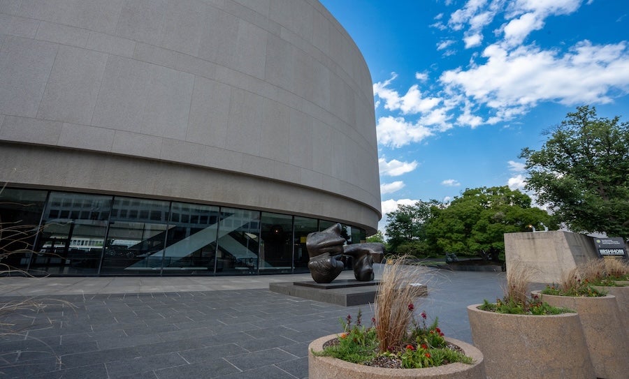 Modern building exterior with curved facade, plaza, plants, and blue sky with clouds