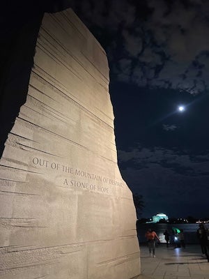 Tall stone wall with engraved text at night under a bright moon, people nearby