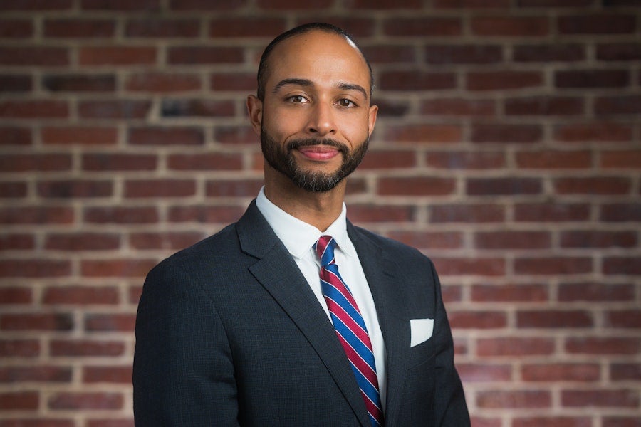 Professional headshot of a person in a dark suit and striped tie, smiling slightly while standing in front of a brick wall background.
