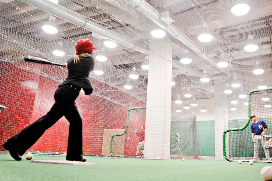 Person swinging a baseball bat in an indoor batting cage while a coach watches from behind a protective net.
