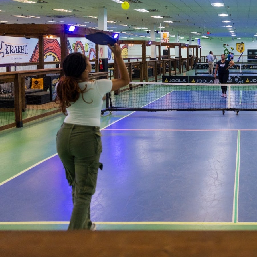 People playing pickleball on an indoor court with a net dividing the playing area and spectators visible in the background.
