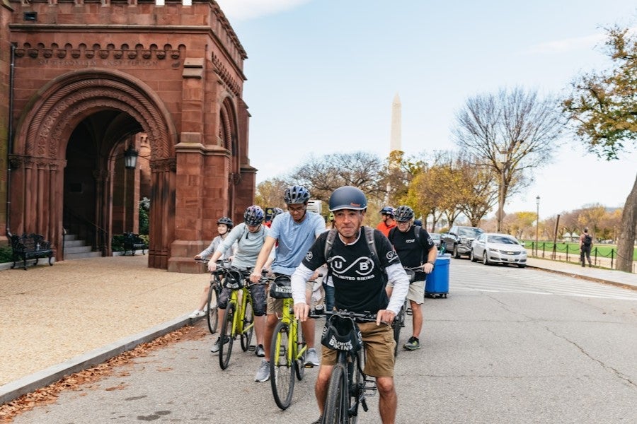 A group of cyclists rides past the Smithsonian Castle on the National Mall, with the Washington Monument visible in the background.