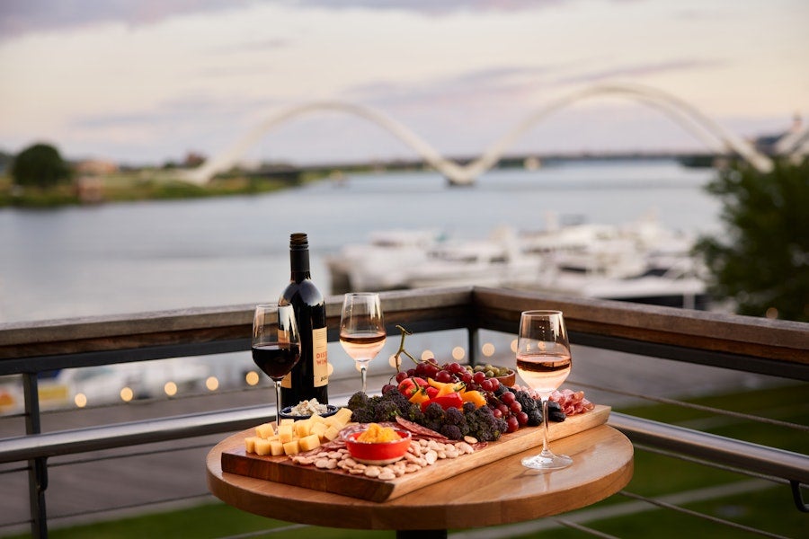 A charcuterie board and glasses of wine sit on an outdoor terrace overlooking a river, marina, and arched bridge at sunset.