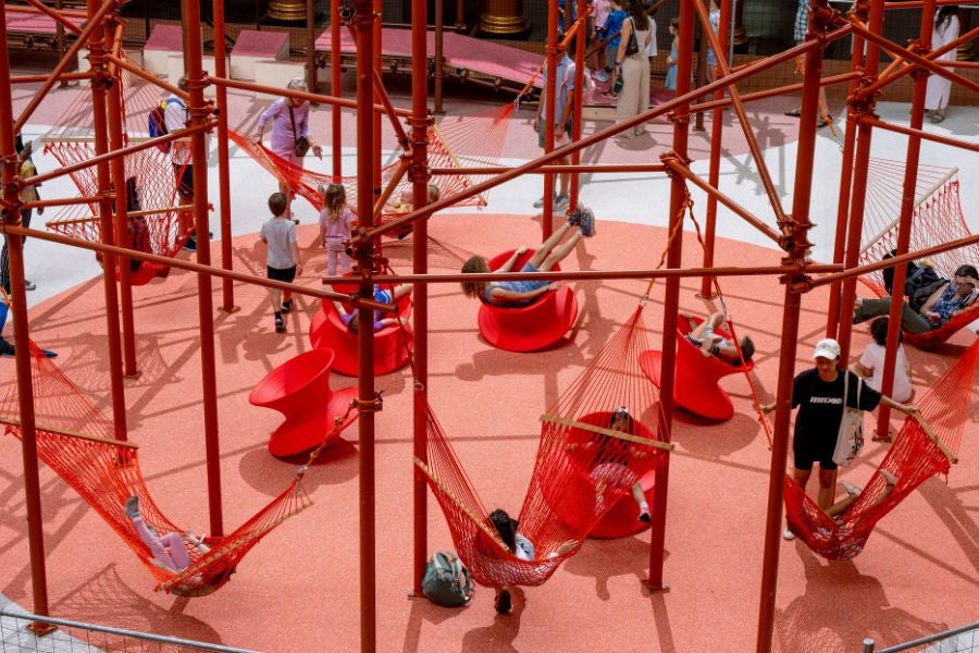 Visitors relaxing in red hammocks suspended within a climbing structure at the National Building Museum’s PLAYGROUND exhibition.