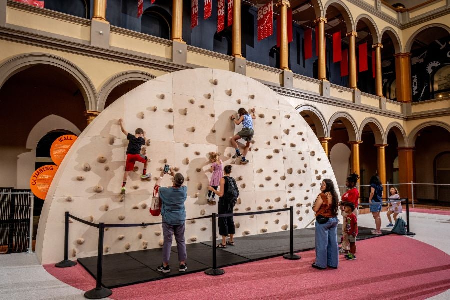 Children climbing an indoor rock wall while families watch at the National Building Museum’s PLAYGROUND exhibition.