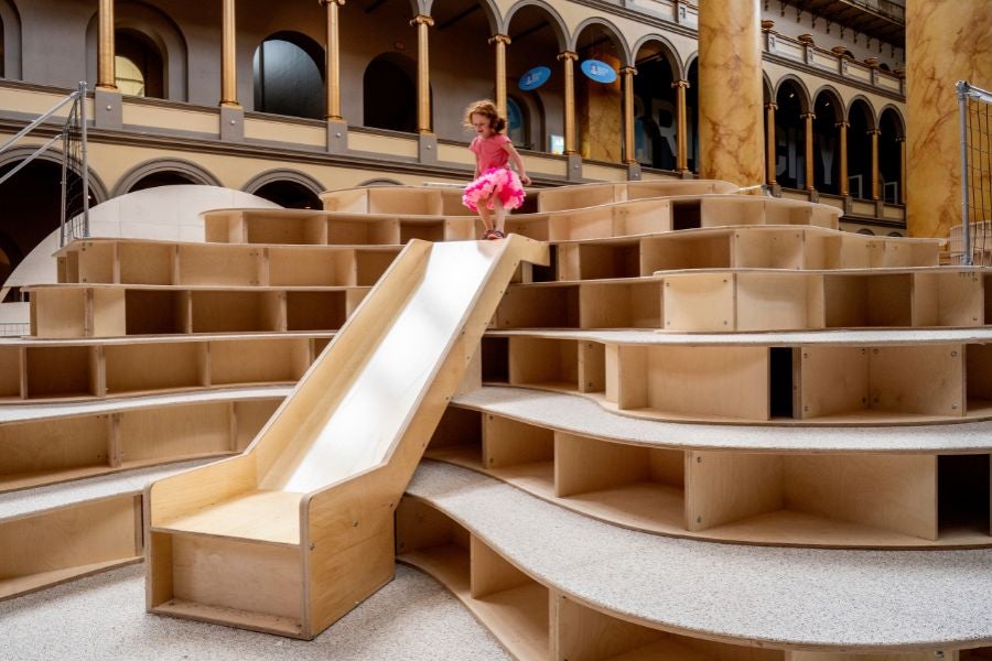 Child sliding down a wooden slide built into a sculptural climbing structure at the National Building Museum’s PLAYGROUND exhibition.