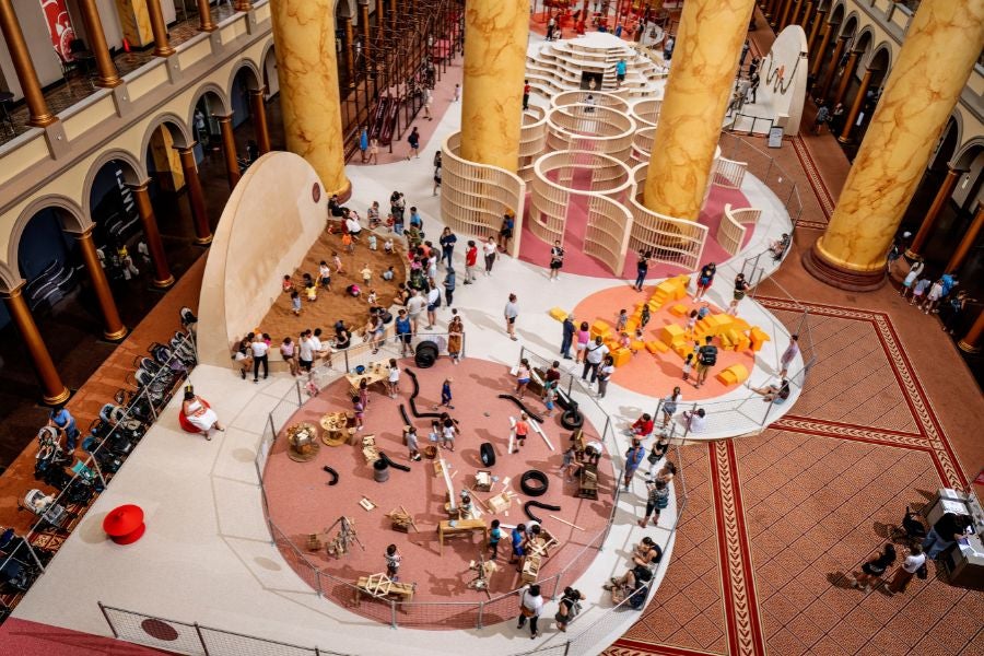 Overhead view of the National Building Museum’s PLAYGROUND exhibition, featuring hands-on building stations, a sand play area, foam blocks and a wooden maze.