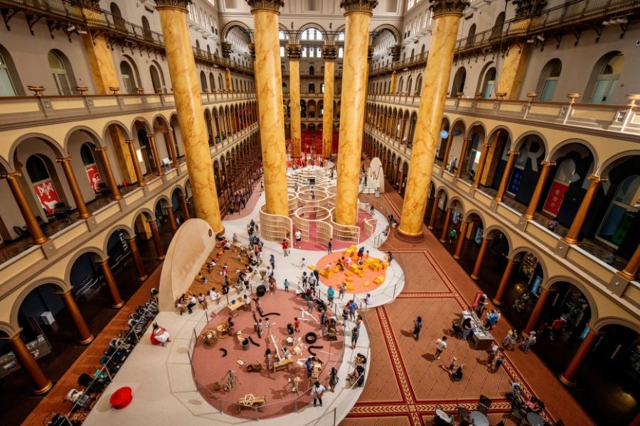 Overhead view of the National Building Museum’s Great Hall filled with visitors exploring the interactive PLAYGROUND exhibition and its colorful play zones.