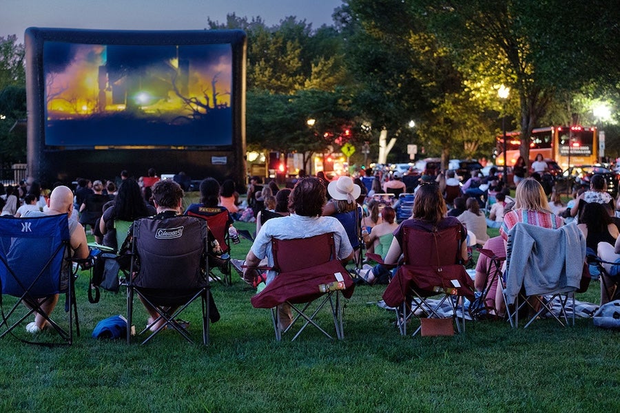 People on lawn chairs watching an outdoor movie at dusk, large screen on the left