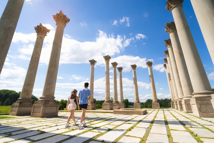 Photo of two people walking among tall stone columns under a blue sky.