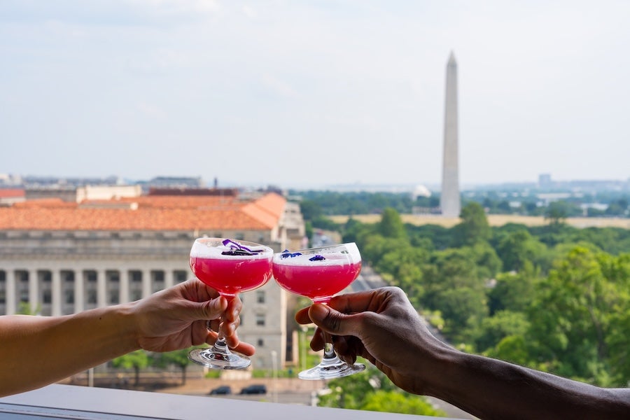 Two hands toast red cocktails with the Washington Monument in the distance