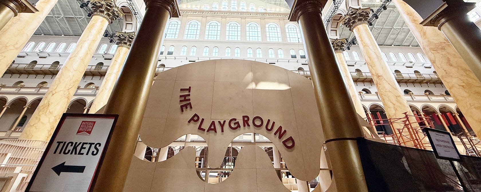 The National Building Museum's Great Hall with tall columns and a banner for its summer installation called “THE PLAYGROUND”