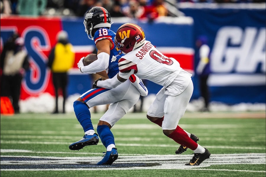 Commanders football player in white tackles Dallas Cowboys ball carrier in blue on green turf