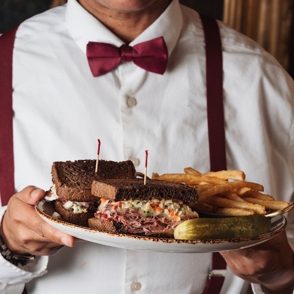 Person in bow tie holds platter of barbecue, fries, pickles and bread slices