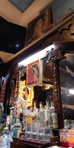 Bar shelf with liquor bottles and glassware under warm lighting, religious icons on wall