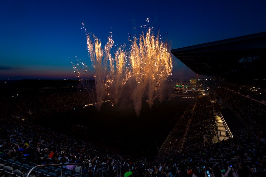 Photo of fireworks bursting above a stadium crowd at dusk